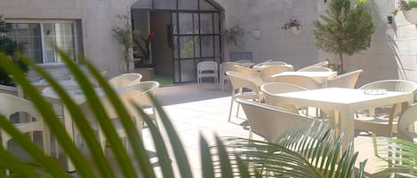 An outdoor seating area with wicker tables and chairs, a cafe entrance, and potted plants.