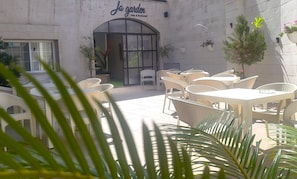 Un espace extérieur aménagé avec des tables et des chaises en osier, l’entrée d’un café et des plantes en pot.
