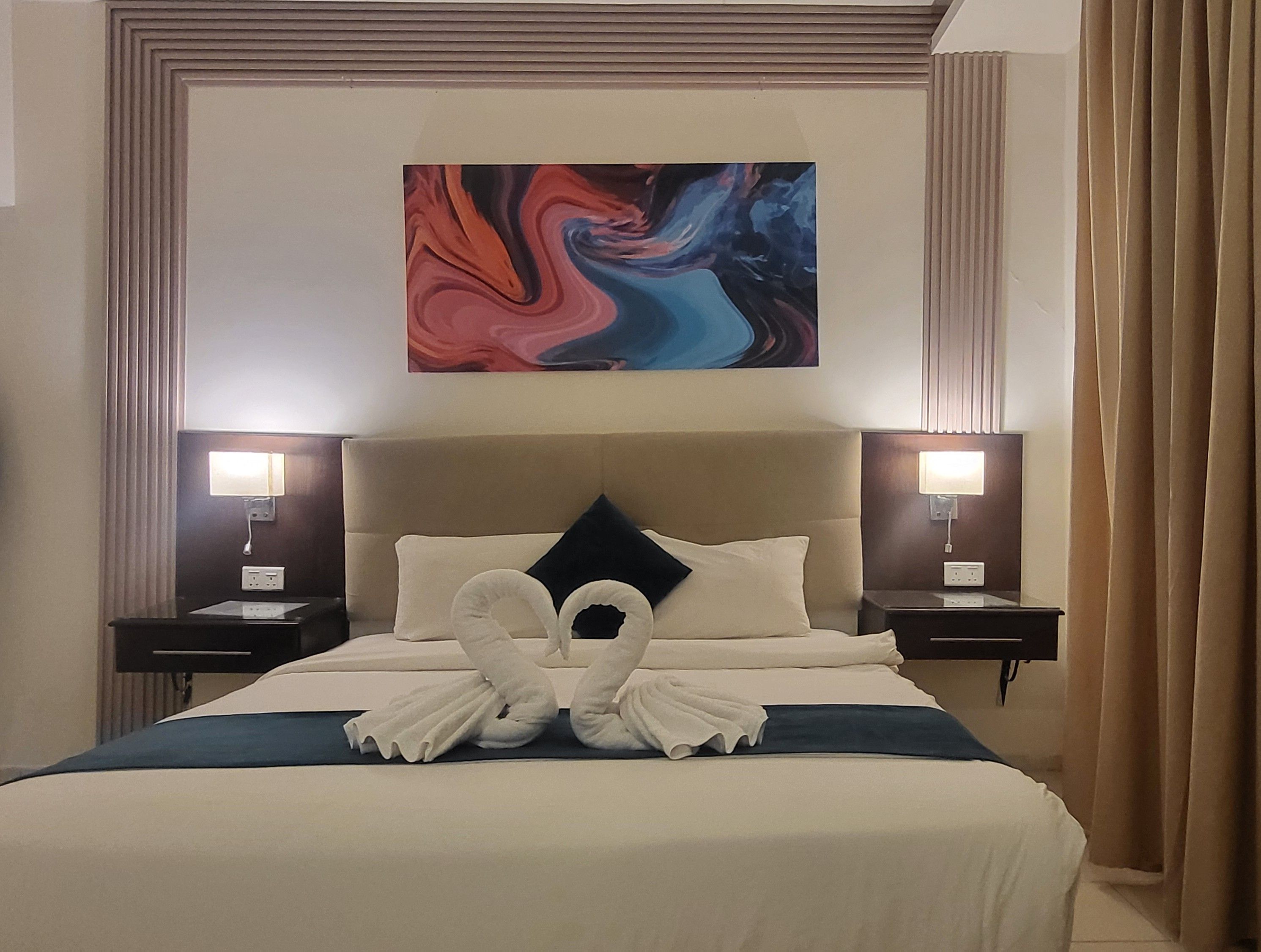 Una habitación de hotel con una cama grande, dos mesitas de noche, un cabecero y un cuadro abstracto colgado en la pared.