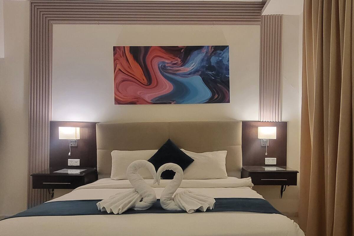 Una habitación de hotel con una cama grande, dos mesitas de noche, un cabecero y un cuadro abstracto colgado en la pared.