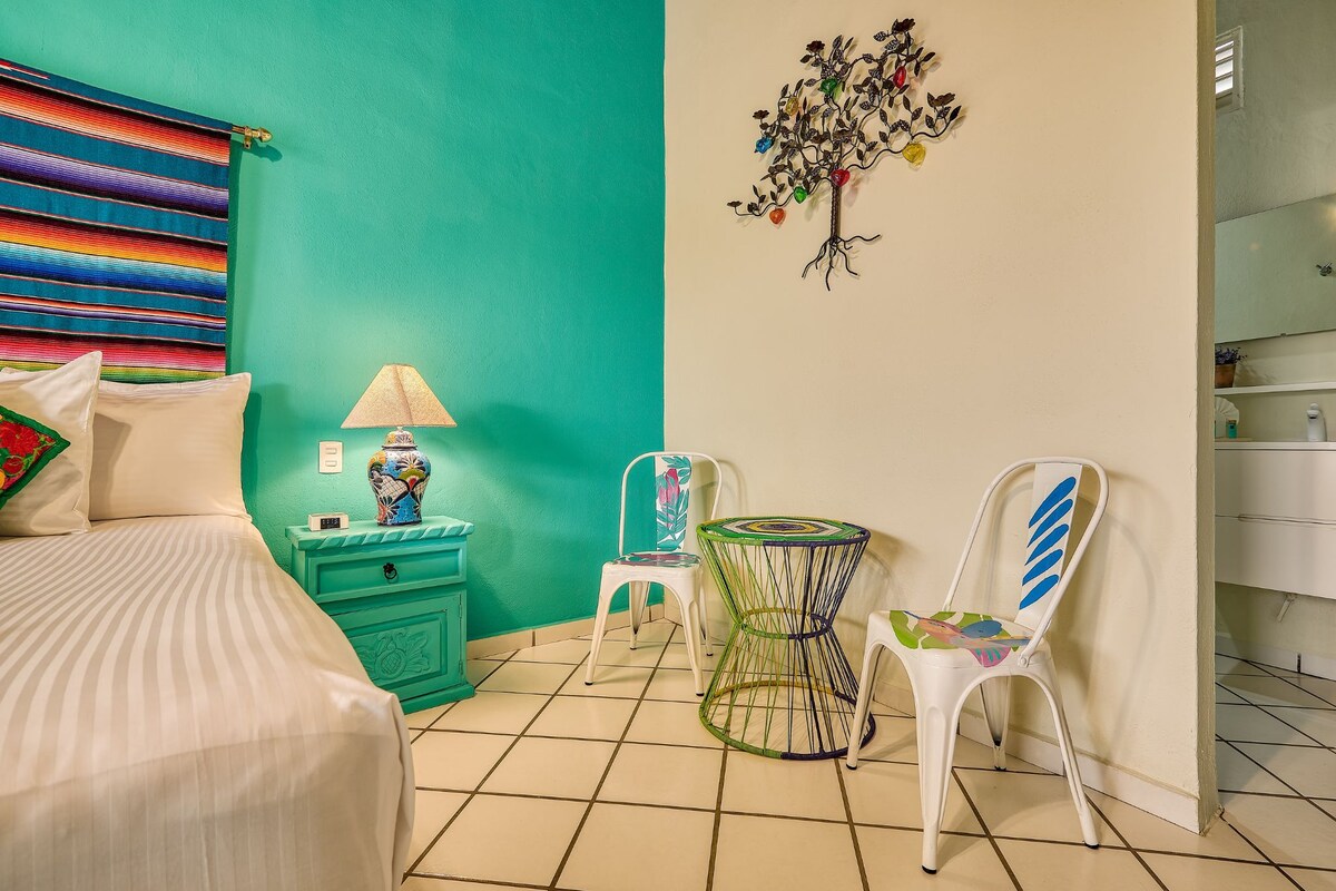 A bedroom with a bed, a nightstand, a lamp, a chair, a small table, and a wall decoration.