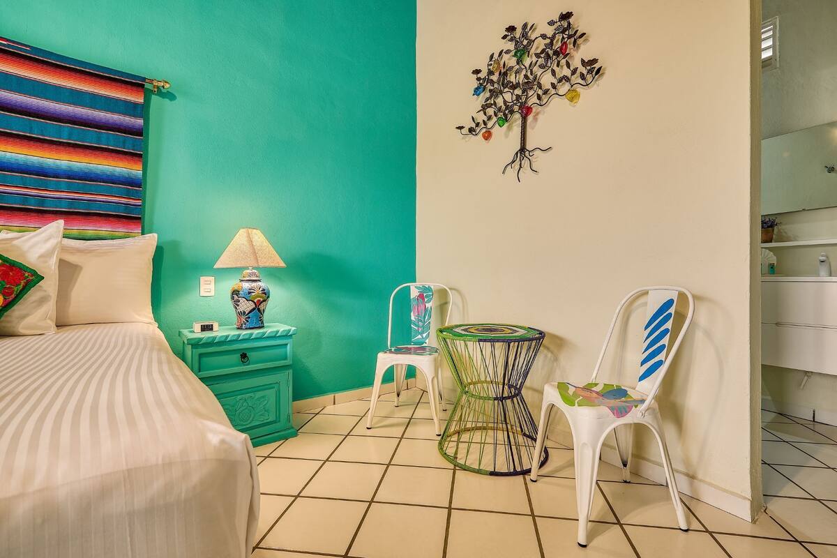 A bedroom with a bed, a nightstand, a lamp, a chair, a small table, and a wall decoration.