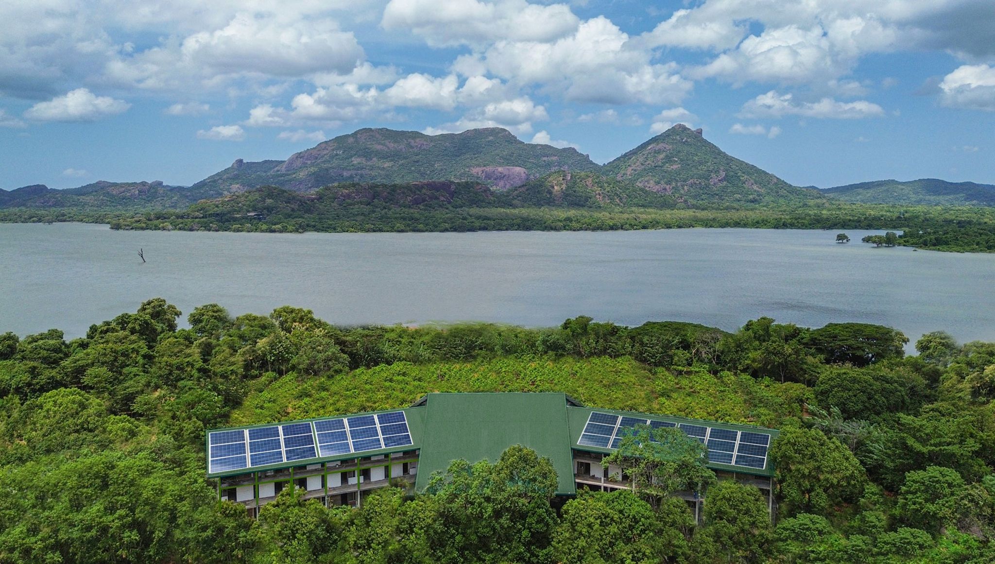 Un edificio con paneles solares en el techo, rodeado de abundante vegetación y un extenso cuerpo de agua con montañas al fondo.
