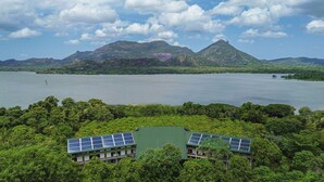 A building with solar panels on the roof, surrounded by dense greenery and a large body of water with mountains in the background. - Amba Yaalu - Thema Collection (Kandalama)
