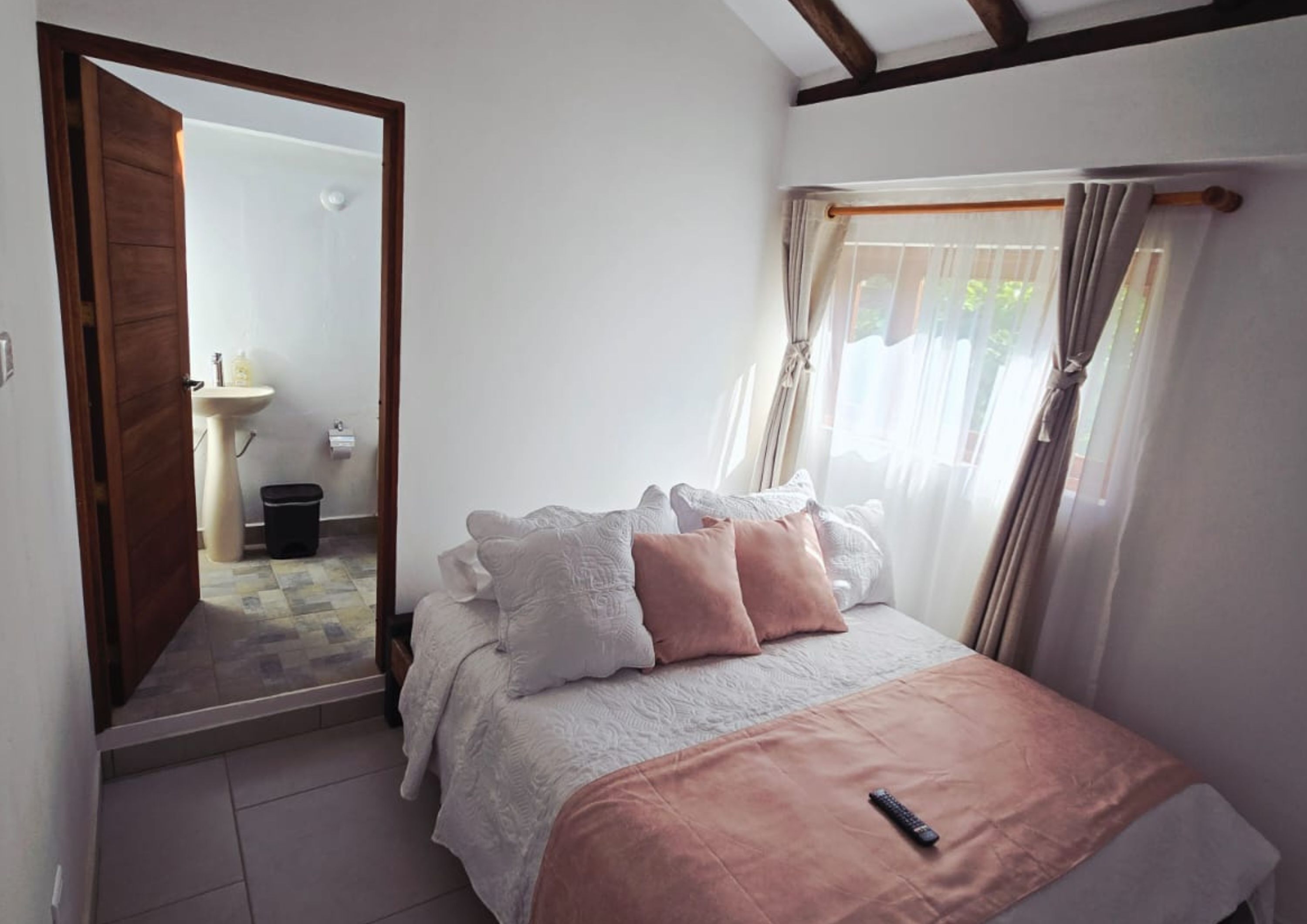 A bedroom with a bed, a remote control, a bathroom visible through an open door, and a window with curtains.