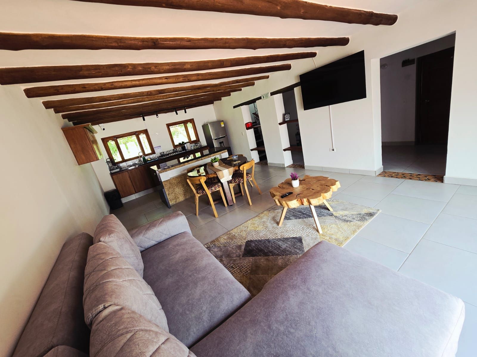 Una sala de estar moderna con un sofá, un comedor con sillas, una cocina al fondo y un televisor de pantalla plana montado en la pared.