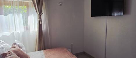 Un dormitorio con una cama, un televisor montado en la pared y una ventana con cortinas.