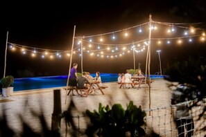 An outdoor dining area with string lights, wooden tables, and a pool in the background. - Adora Bay Ke Ga Beach Resort (Tan Thanh)