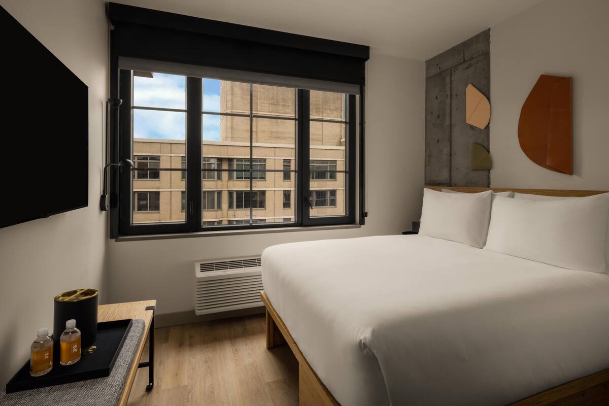 Habitación de hotel con un ventanal grande, una cama, una mesita de noche con botellas y una obra de arte colgada en la pared.