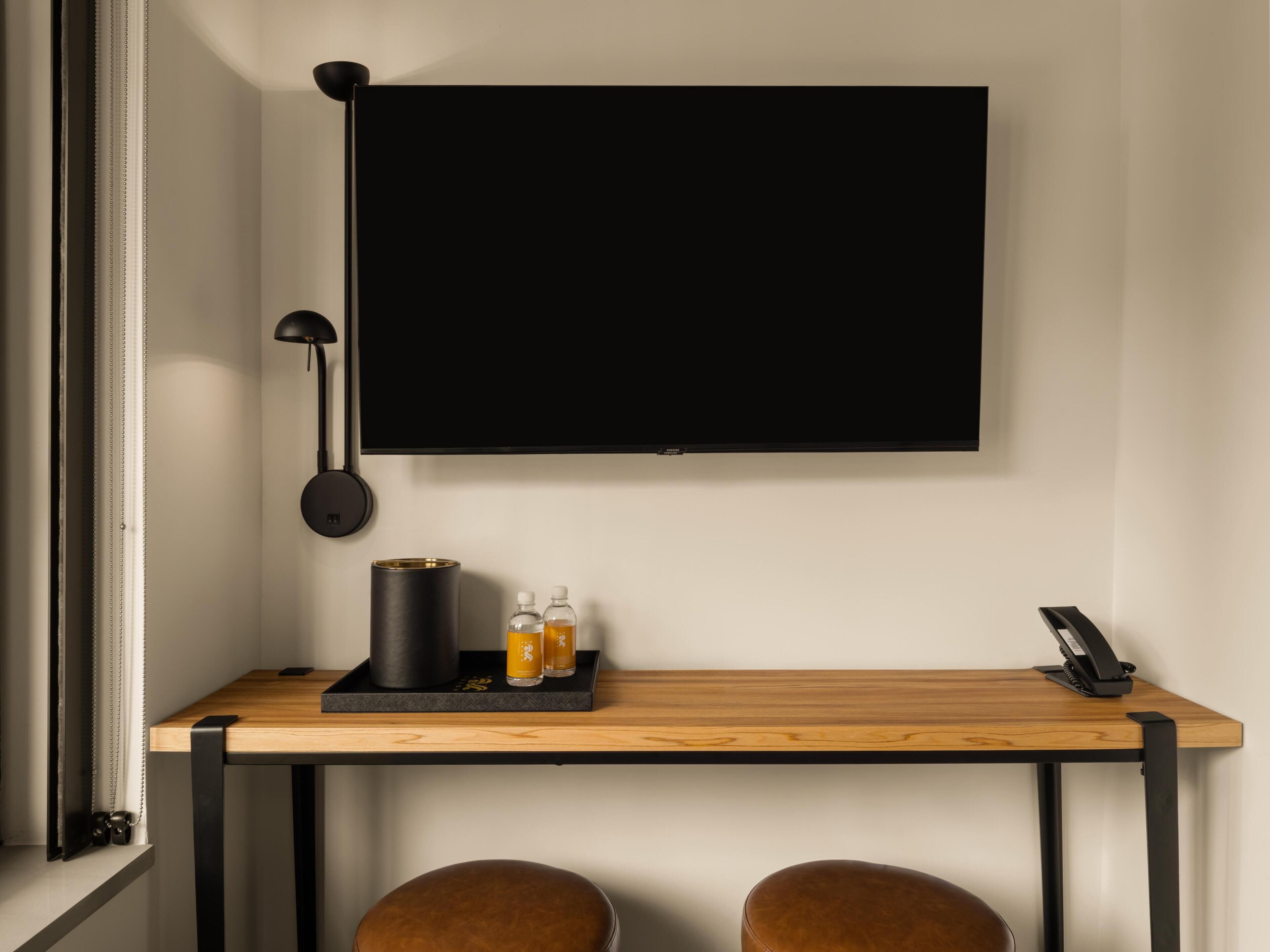 Una habitación moderna con una televisión de pantalla plana grande montada en la pared, un escritorio con lámpara, una cafetera y dos botellas de agua.