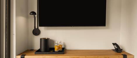 Una habitación moderna con una televisión de pantalla plana grande montada en la pared, un escritorio con lámpara, una cafetera y dos botellas de agua.