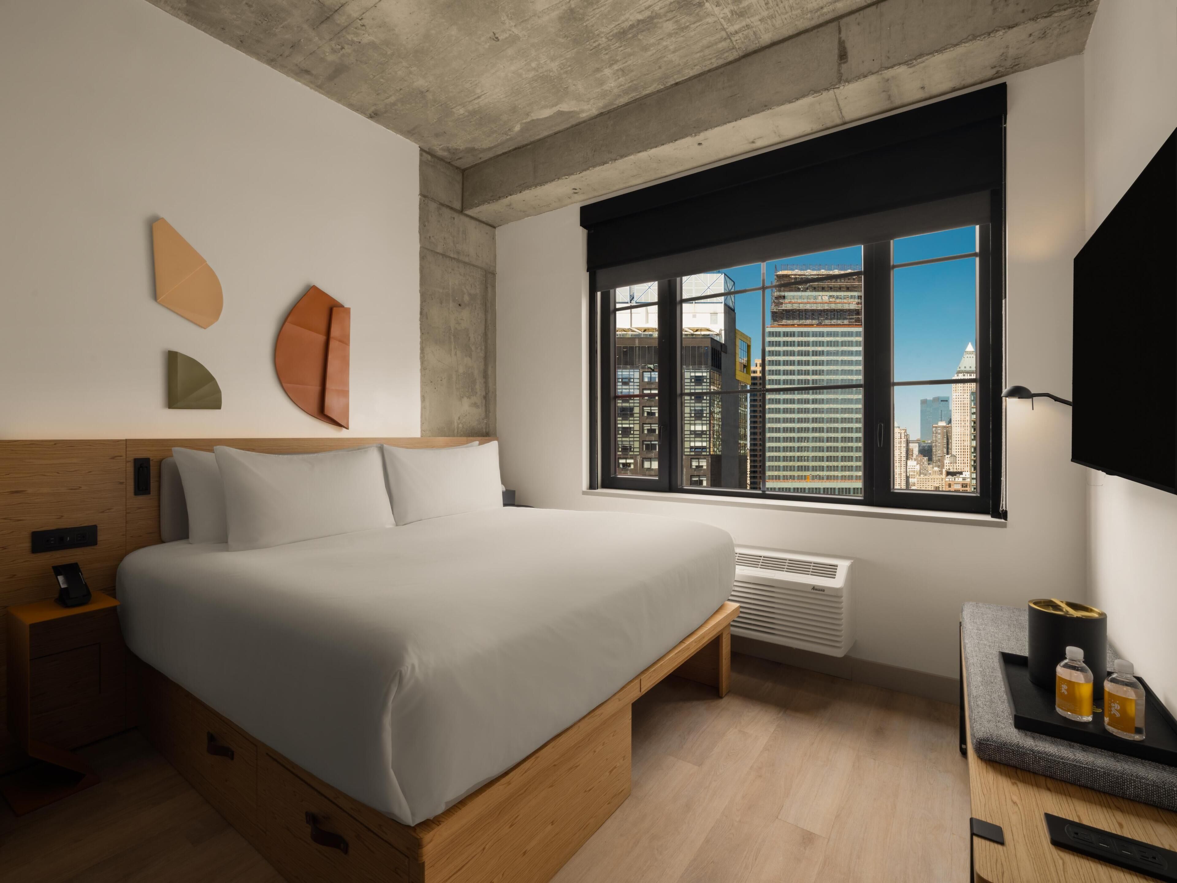 Quarto de hotel moderno com uma cama grande, mesa de cabeceira, escrivaninha com luminária, janela com vista para a cidade e televisão na parede.