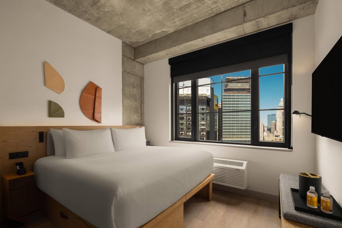 Habitación de hotel moderna con una cama grande, una mesita de noche, un escritorio con lámpara, un ventanal con vistas a la ciudad y un televisor montado.