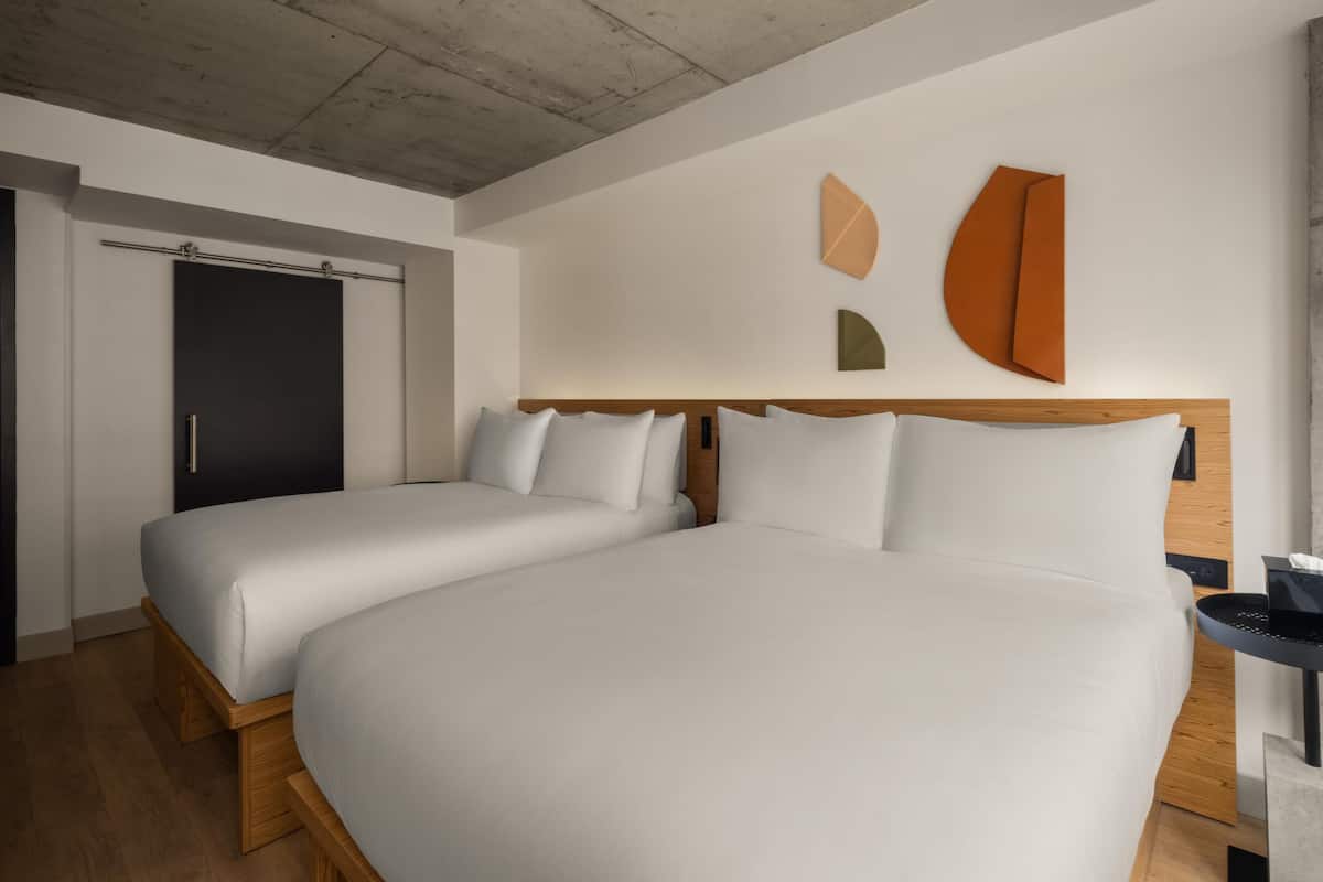 Una habitación de hotel moderna con dos camas, un cabecero de madera, un techo de hormigón y una obra de arte abstracto colgada en la pared.