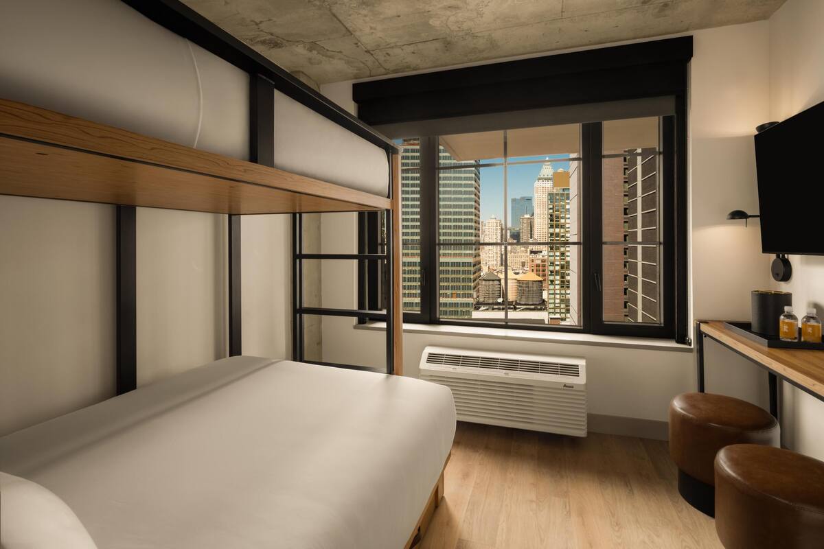 Una habitación de hotel moderna con una cama grande, un ventanal con vistas a la ciudad, un escritorio con lámpara y un radiador.