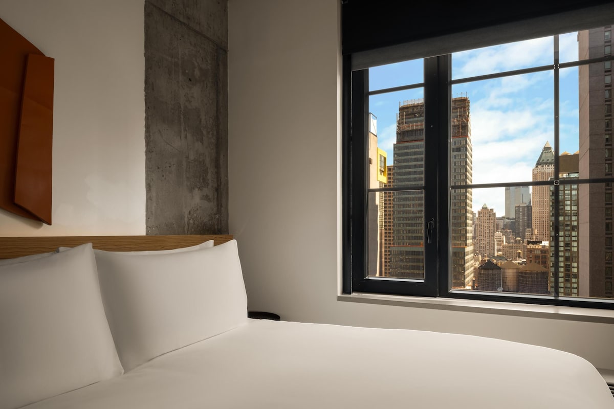 Una habitación de hotel con una cama grande, vista al horizonte de la ciudad a través de una ventana y una pared de concreto.