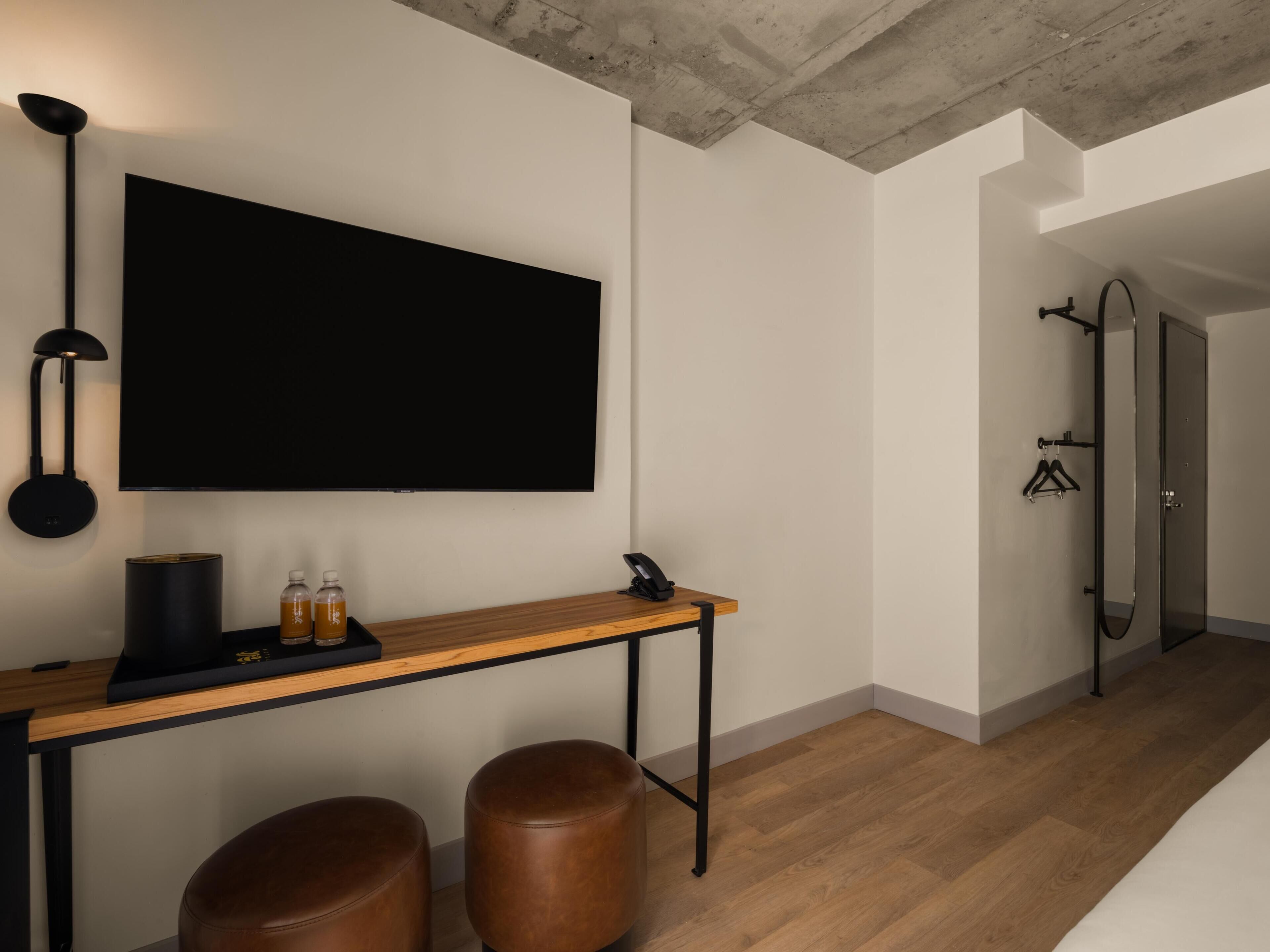 Une chambre moderne dotée d’un grand téléviseur à écran plat, d’un bureau en bois, de deux tabourets en cuir, d’une lampe suspendue et d’un miroir.