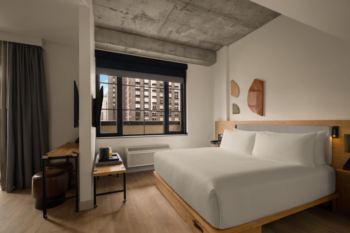 Una habitación de hotel moderna con una cama grande, un escritorio, una silla, una ventana con vistas a edificios y un techo con concreto a la vista.