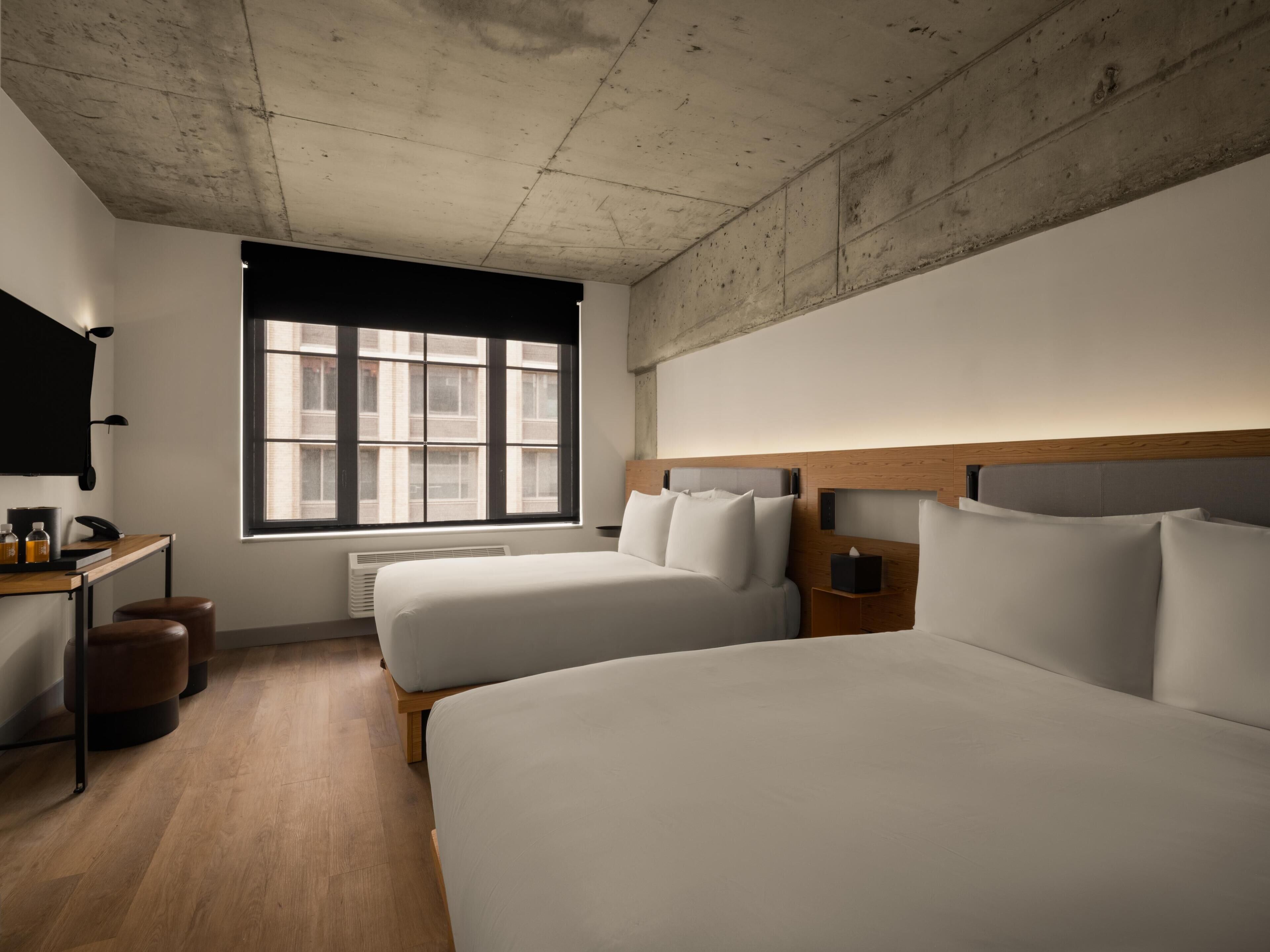 Quarto de hotel com duas camas, uma escrivaninha, uma cadeira, televisão e janela com vista para edifícios.