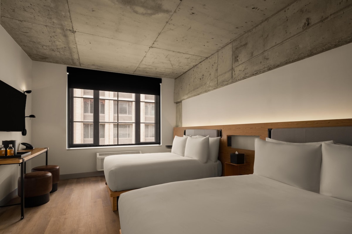 Habitación de hotel con dos camas, un escritorio, una silla, un televisor y una ventana con vista a edificios.