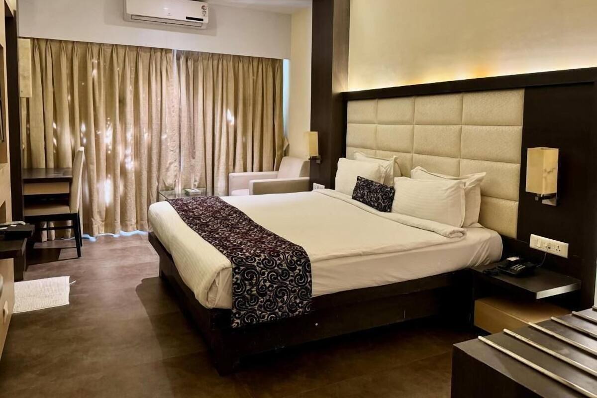 A hotel room with a large bed, a desk, a chair, a nightstand, and a window with curtains.