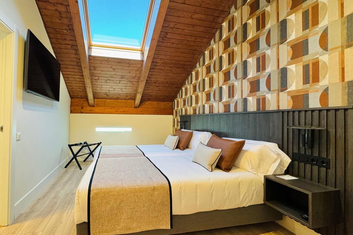 Un dormitorio con techo inclinado, una cama con almohadas, un televisor montado en la pared y un diseño de pared estampado.