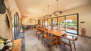 A spacious dining area with a long wooden table and chairs, large windows, and a view of the outdoor patio. - Cabana Kedungu Villa By Ini Vie Hospitality (Payangan)