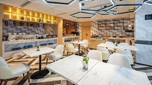 A modern restaurant with marble flooring, white tables, and chairs. There are hexagonal ceiling lights and a kitchen area with a buffet. - Mercure Shanghai Pudong Zhangjiang Park (Zhoushan)