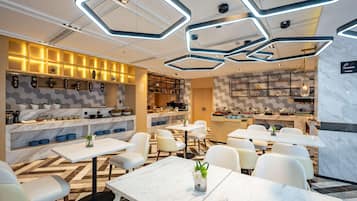 Restaurante moderno com piso de mármore, mesas e cadeiras brancas. Apresenta luminárias hexagonais no teto e uma área de cozinha com buffet.