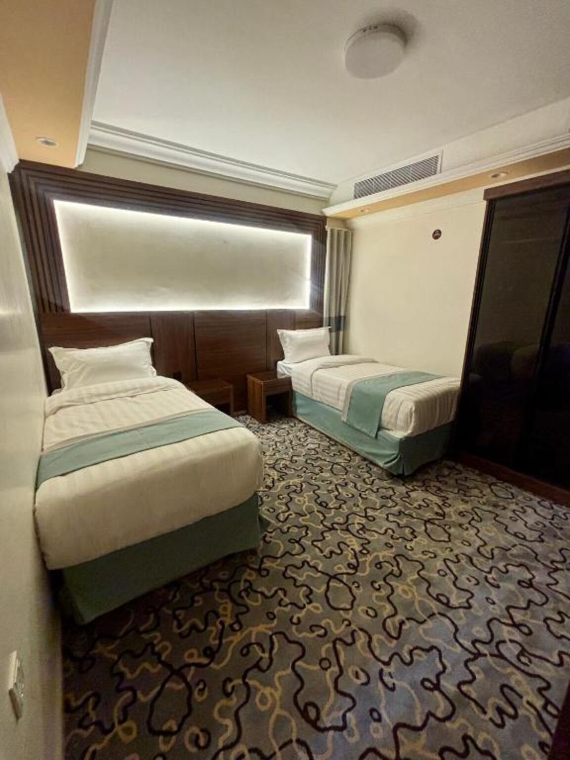 A hotel room with two beds, a patterned carpet, a large window with blinds, and a ceiling vent.