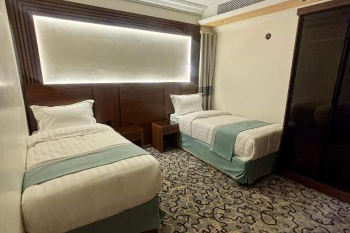 A hotel room with two beds, a patterned carpet, a large window with blinds, and a ceiling vent.
