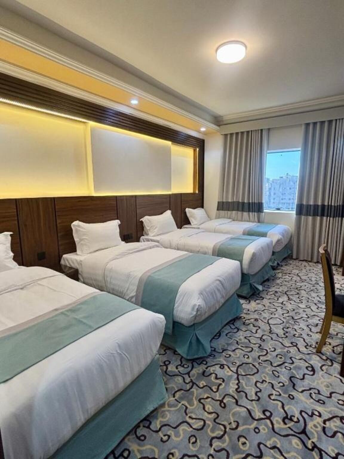 A hotel room with four beds, a patterned carpet, a wooden headboard, and a window with curtains.