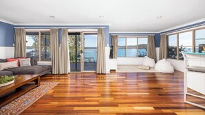 Ein geräumiges Wohnzimmer mit Holzdielenboden, einer grauen Couch, einem weißen Sitzsack und großen Fenstern mit Meerblick.