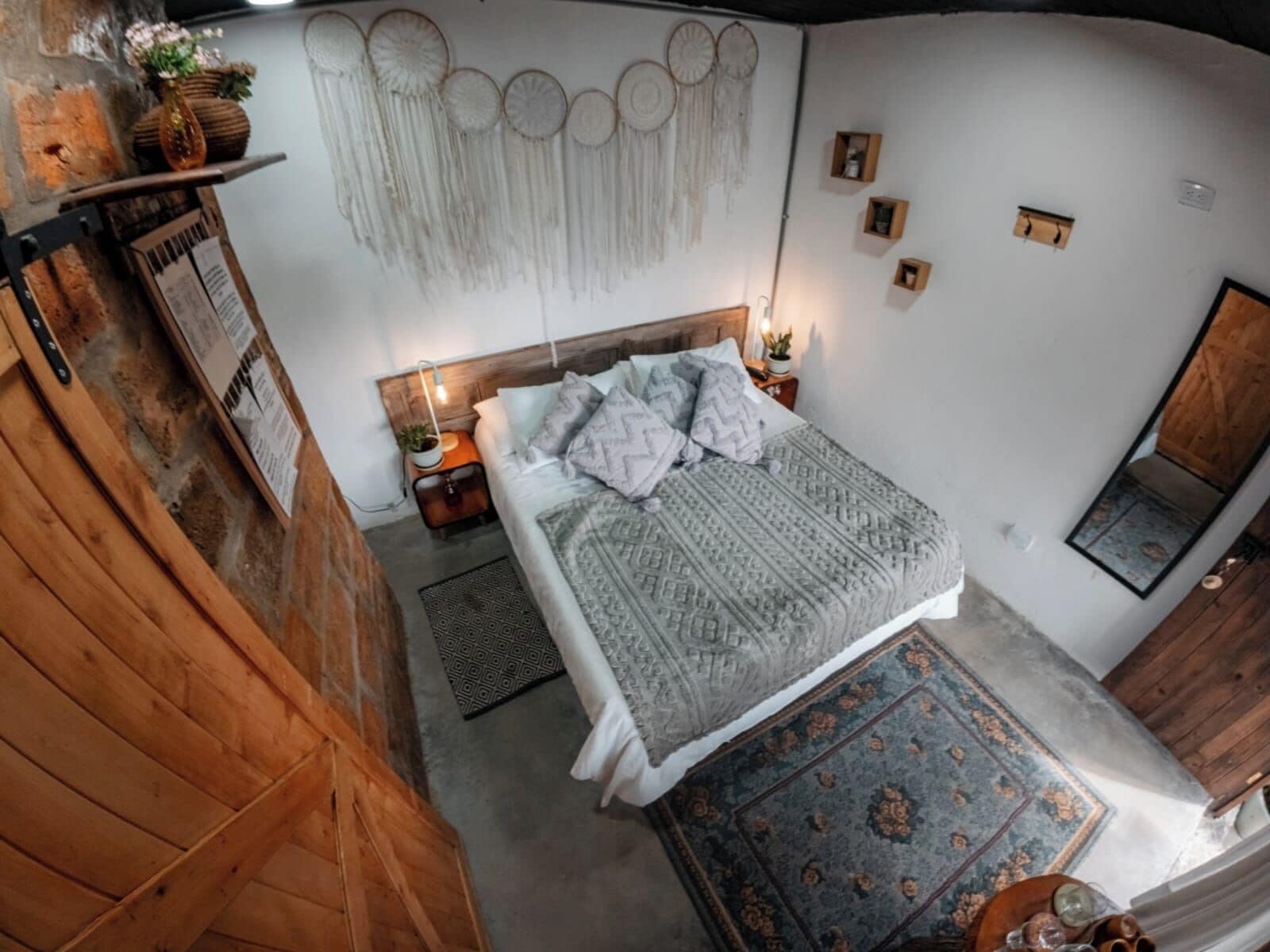 A bedroom with a wooden headboard, a patterned bedspread, a decorative wall hanging, a small bedside table with a lamp, and a mirror.