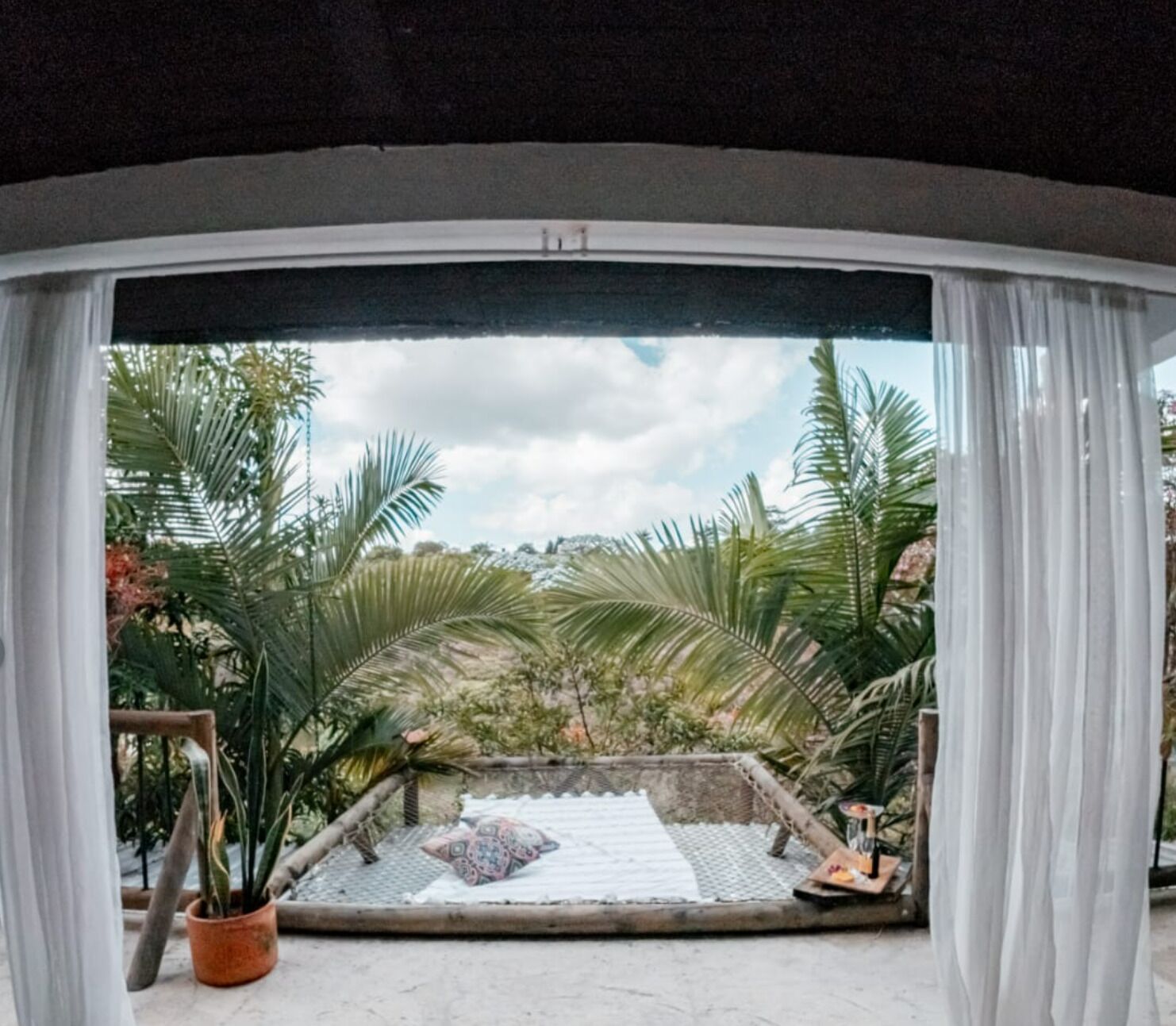 A view through a window with white curtains, revealing a patio area with a patterned rug, a small table with a candle, and various potted plants.