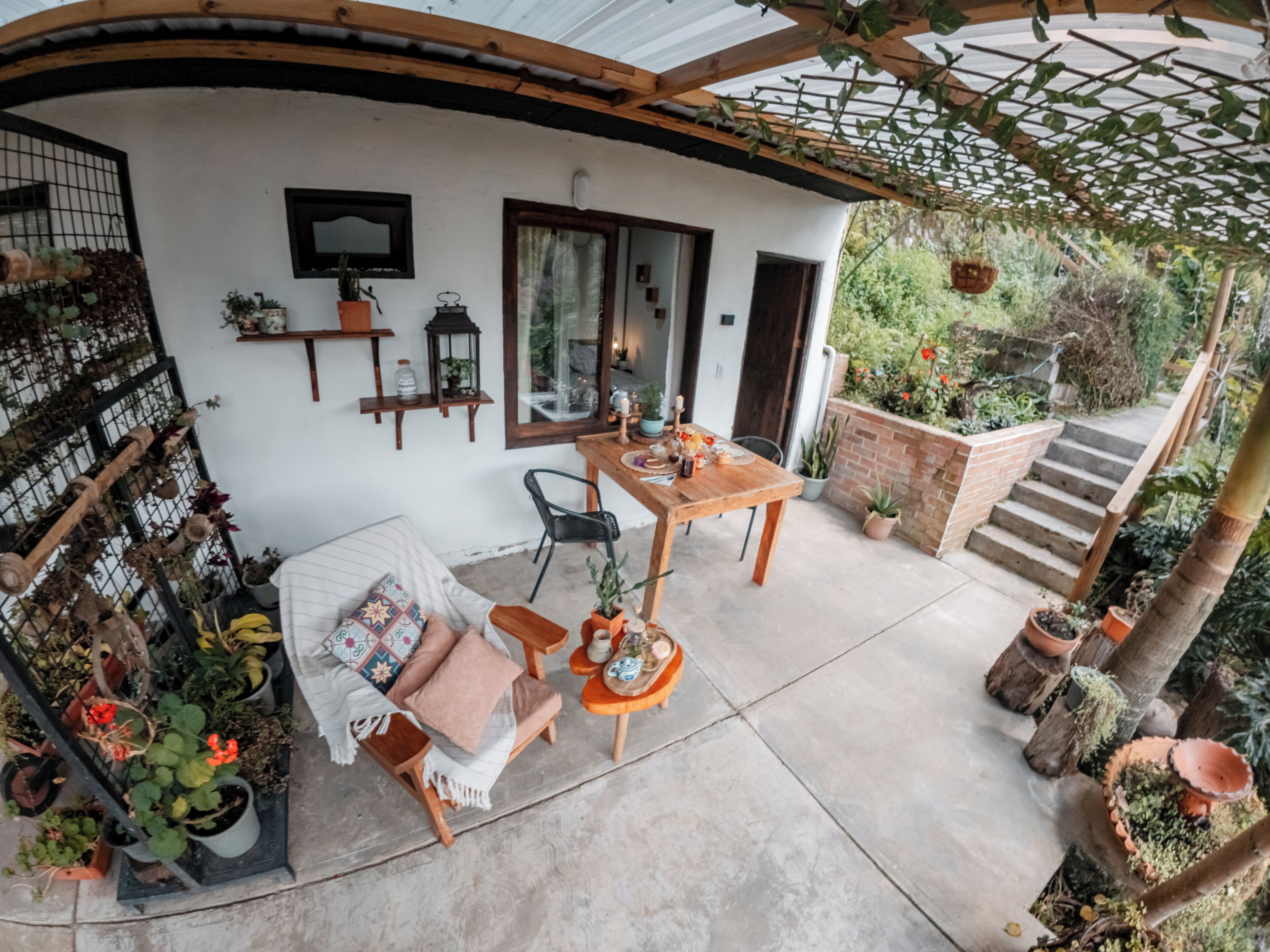 A covered patio area with a wooden table and chairs, surrounded by potted plants and a small seating area.