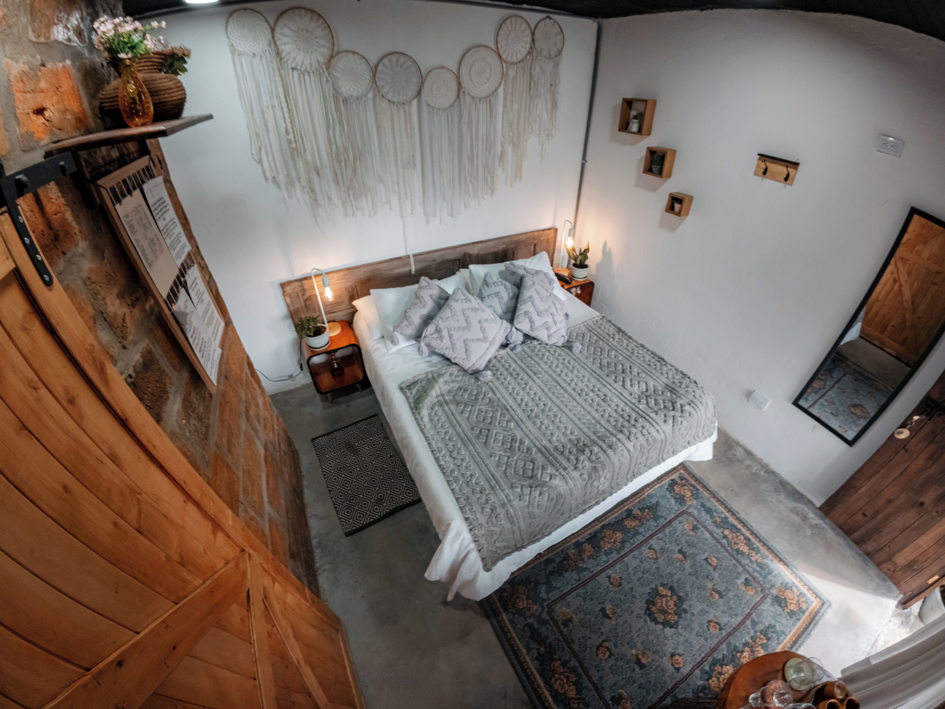 Un dormitorio con cabecera de madera, cama con ropa de cama estampada, mesita de noche con lámpara, espejo adosado a la pared y un adorno colgante en la pared.