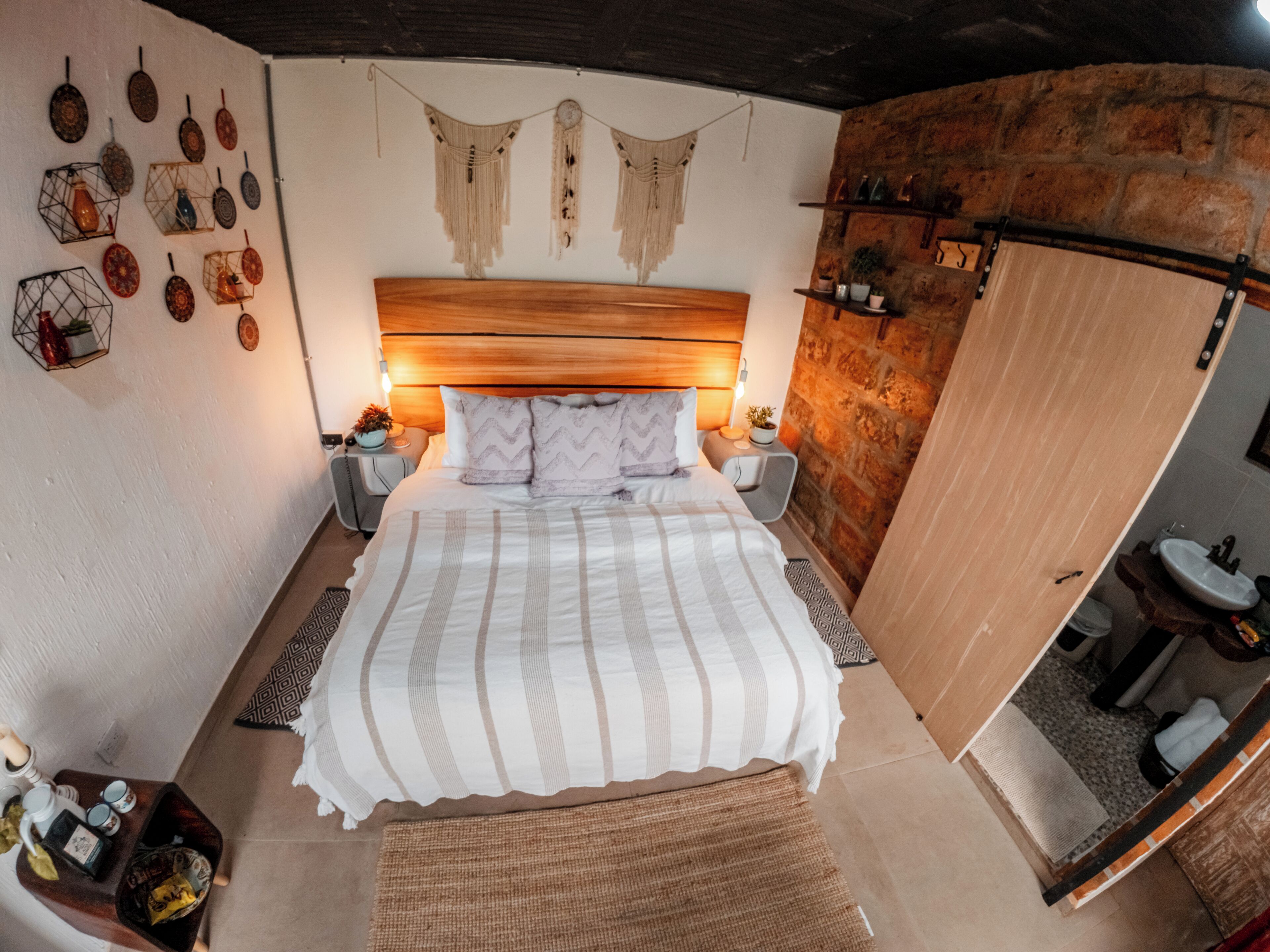 A bedroom with a wooden headboard, a bed with striped bedding, bedside tables with lamps, a wall with hanging decorations, and a bathroom with a sink and mirror.