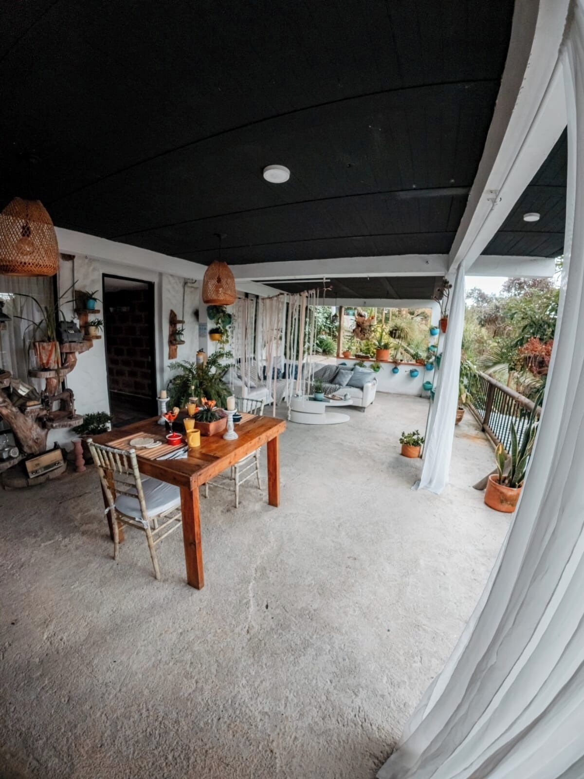 An outdoor patio with a wooden table and chairs, surrounded by potted plants and a white curtain.