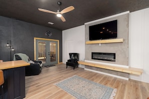 A modern living room with a wooden floor, a ceiling fan, a flat-screen TV mounted on a wall, a fireplace, and a seating area with a chair and a sofa. - Peaceful, NEWLY Constructed Barndominium Retreat Near Fort Leonard Wood (Waynesville)