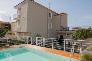 A swimming pool with a wooden deck, a white railing, and a building with balconies and a red-tiled roof. - Dama Luxury Apartment (Pompei)