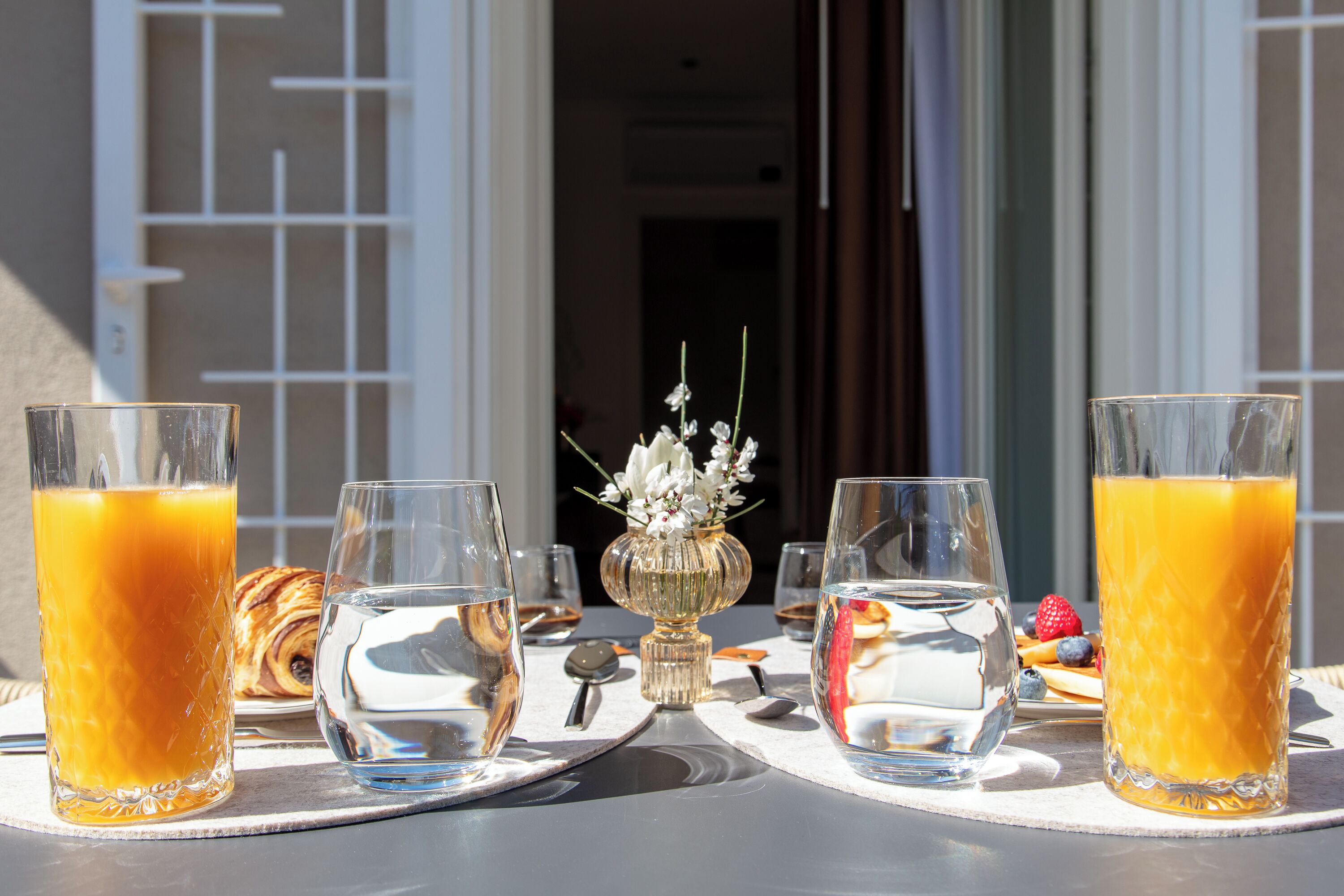 A breakfast table with two glasses of orange juice, a water glass, a croissant, and a small vase with flowers.