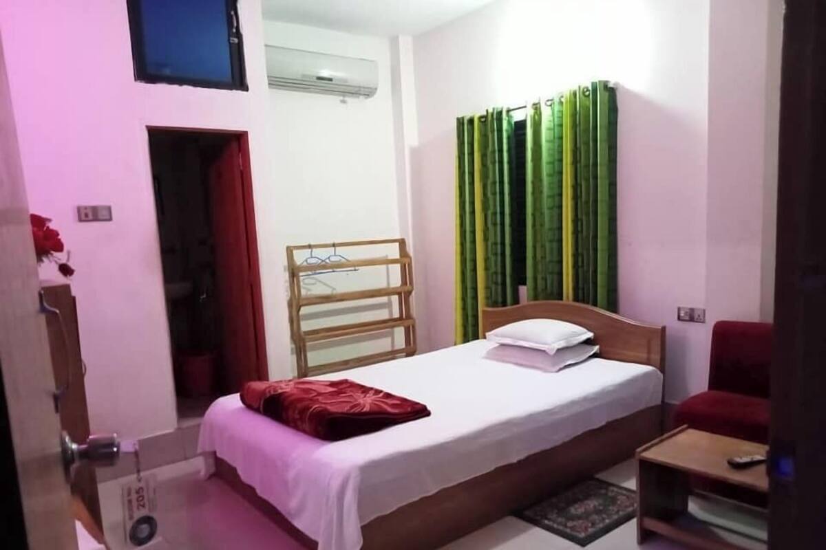 A bedroom with a bed, a wooden headboard, a small bedside table, a red chair, and a window with green curtains.