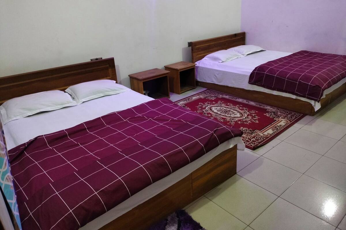 A room with two single beds, each with a wooden headboard and a red bedspread. There is a small wooden nightstand between the beds, and a patterned rug on the floor.