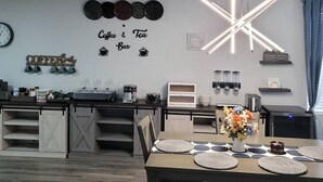 A modern kitchen with a dining area, featuring a coffee and tea bar, a unique ceiling light, and a clock on the wall.
