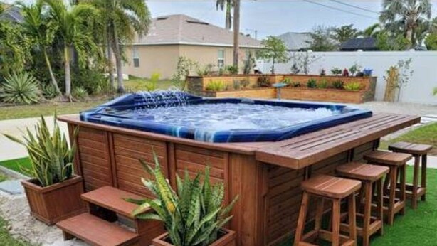 A hot tub with wooden steps and bar stools, surrounded by potted plants and a grassy area.