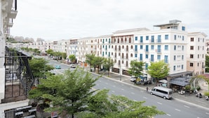 Une rangée de bâtiments blancs aux fenêtres et balcons bleus, une rue avec des voitures et un fourgon, et des arbres bordant le trottoir.