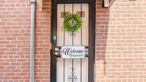A brick building with a black door, a 'Welcome Guest' sign, and a small welcome mat.