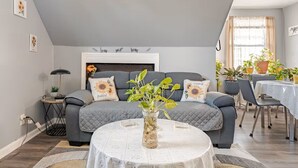 A living room with a grey sofa, a round coffee table, and a fireplace. There are potted plants on the windowsill and a dining table with chairs.