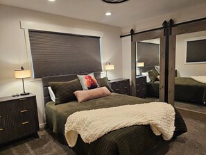 A bedroom with a bed, bedside tables, a mirror, and a window with blinds. - Summer and Winter Getaway (Sandy)